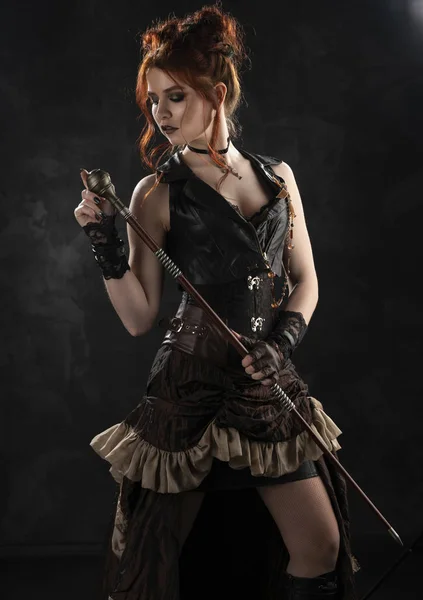 Düşünceli derin bir yaka ile büyük bir meme Victorian tarzı steampunk kostüm giyen bir güzel kızıl saçlı cosplayer kız bir baston ellerinin karanlık bir arka plan üzerinde tutar. Kopya alanı.