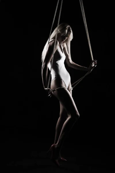 Beautiful slender blonde girl, wearing a white bodisuit and red stilettos, sensually plays and poses with the ropes. Black background. Artistic noir silhouette photo. Copy space.