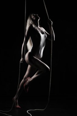 Güzel bir ince sarışın kız, beyaz bodisuit ve kırmızı stilettos, sensually asmak ve halatlar üzerinde dans ediyor. Siyah arka plan. Sanatsal noir siluet fotoğraf. Kopya alanı.