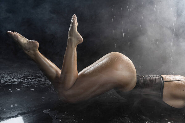 Booty big breasted girl, dressed in a black translucent body sensually lays on the floor under water droplets in theatrical smoke. Close up on her ass. Advertising and commercial design. Copy space.