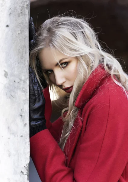 A beautiful blond girl wearing a red coat and gloves stands by a — Stock Photo, Image