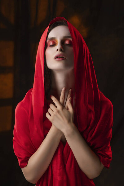 Beautiful girl with fashionable makeup in red tones, wearing a red silk scarf thrown over her head that covers her naked breast, sensually poses on the background of the shadow from the window.