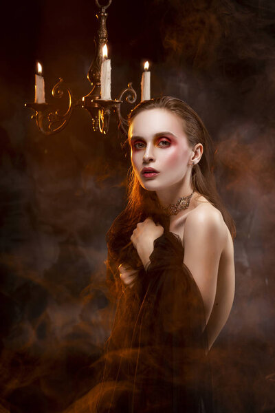 A beautiful slim topless girl, wearing earrings and a necklace, and covering her nudity with a black veil, sensually poses in theatrical smoke near the chandelier with lightning candles