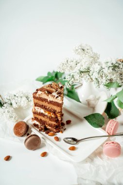 Cam bir tabakta çeşitli kurabiye ve fındıkla süslenmiş çikolatalı pasta ve leylak ve yeşil yapraklardan oluşan beyaz çiçekler arasında badem kurabiyesi. Yemek fotoğrafçılığı. Reklamcılık ve ticari yakın plan tasarımı
