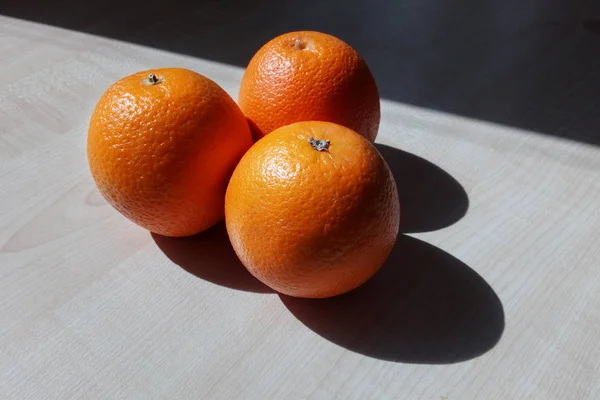 三只橘子躺在一张木桌上 用一个三角形的形状照亮了明亮的清晨的太阳 有锐利的阴影 — 图库照片