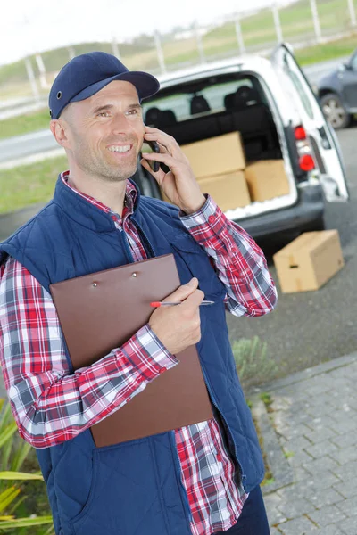 En bild av en glad deliveryman hålla låda och mobiltelefon — Stockfoto