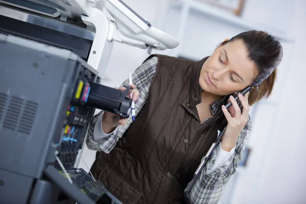 Νεαρή γυναίκα επισκευαστή σχετικά με το τηλέφωνο που στέκεται δίπλα στον εκτυπωτή — Φωτογραφία Αρχείου