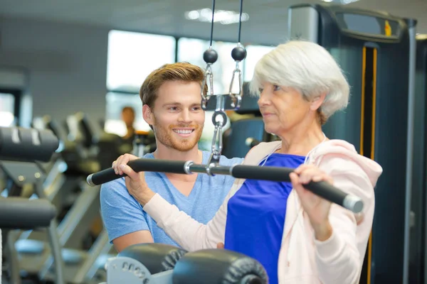 Mujer mayor haciendo ejercicio en la máquina de resistencia con entrenador personal —  Fotos de Stock