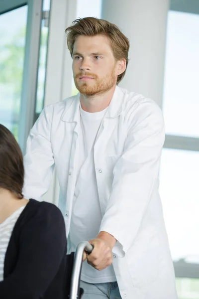 Hübsche junge Krankenschwester schubst Patientin im Rollstuhl — Stockfoto