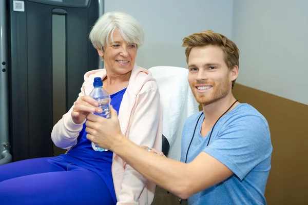 Kvinnliga senior med vattenflaska och tränare på gymmet — Stockfoto