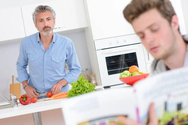 Zwei Männer in der Küche, einer bereitet Gemüse zu, der andere liest aus einem Rezeptbuch — Stockfoto