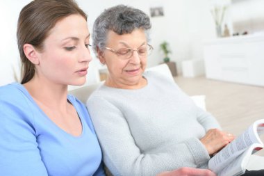 young female caregiver reading a story to the old woman clipart