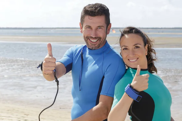 Paar dragen wetsuits op het strand waardoor duim omhoog gebaren — Stockfoto