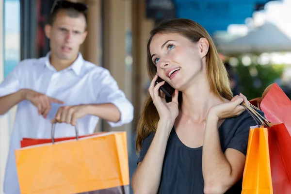 Молодая женщина с телефоном и покупками, пока ее парень ждет — стоковое фото