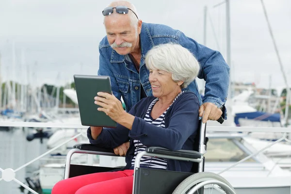 Pareja de ancianos en el puerto mirando a la mujer tableta en silla de ruedas — Foto de Stock
