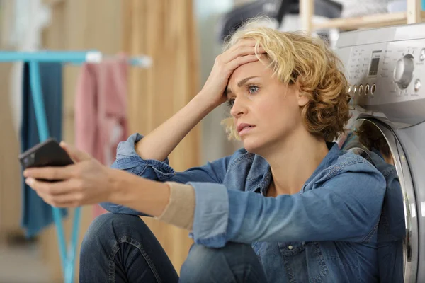 Mujer angustiada sosteniendo teléfono inteligente apoyado contra la lavadora — Foto de Stock