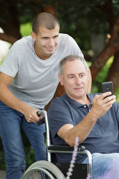 Hijo con silla de ruedas padre tomando selfie en parque — Foto de Stock