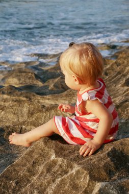 Harcama zaman açık havada küçük olanlar için önemli olan kayalık kıyısında oturan kırmızı ve beyaz elbiseli bir yaşındaki bebek kız