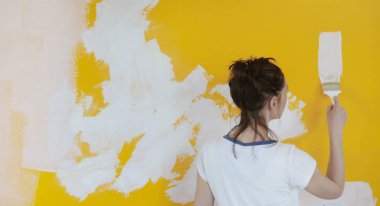 Sarı bir duvarın üzerinde beyaz boyayla resim yapan genç bir kadın.