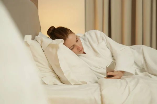 若い女性がベッドに横たわって電話でメッセージをチェック — ストック写真