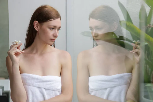 一个美丽的年轻女子在浴室的肖像 穿着白色毛巾 拿着弗兰吉帕尼花 放松和身体护理的概念 — 图库照片