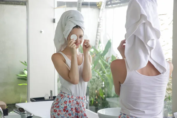 バスルームに立って頭の上にタオルを持つ美しい若い女性の肖像画と鏡の前に綿のパッドで彼女の顔を掃除 メイクを削除 美しさとスパの概念 — ストック写真