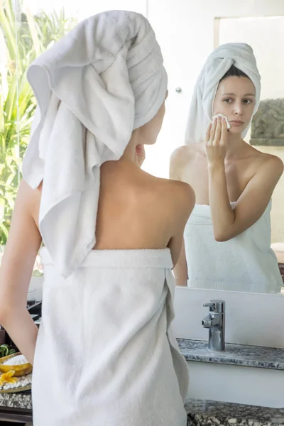 バスルームに立って頭の上にタオルを持つ美しい若い女性の肖像画と鏡の前に綿のパッドで彼女の顔を掃除 メイクを削除 美しさとスパの概念 — ストック写真