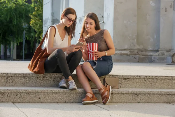 夏の日に通りの階段に座って ポップコーンを食べて 携帯電話を見て 2人の女の子が街でぶらぶらしている — ストック写真
