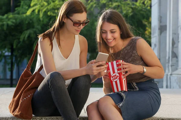两个朋友坐在街楼梯上 吃着爆米花 看着手机 两个女孩在城市里闲逛 — 图库照片
