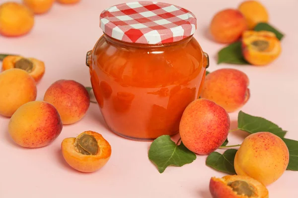 Jar of apricots jam and fresh apricots on pink background