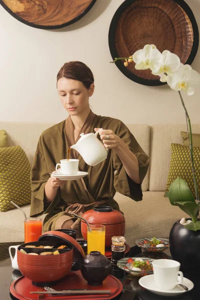 Woman in robe pouring a coffee or tea in the cup before having a big healthy Japanese breakfast