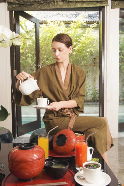 Woman in robe pouring a coffee or tea in the cup before having a big healthy Japanese breakfast