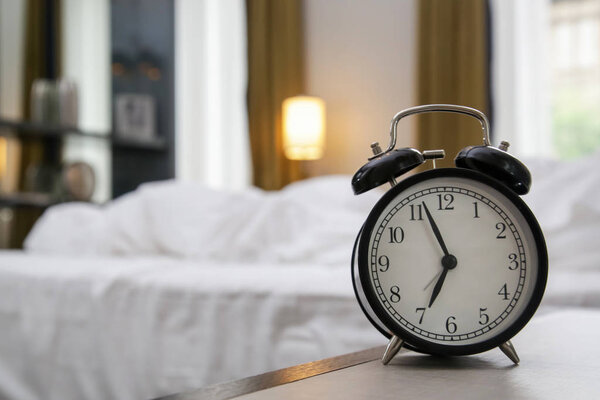 Классический черный будильник на тумбочке показывает семь часов со спальней на заднем плане
