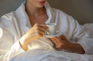 Beyaz bornozla yatakta sabah kahvesi içen genç ve güzel bir kadın.