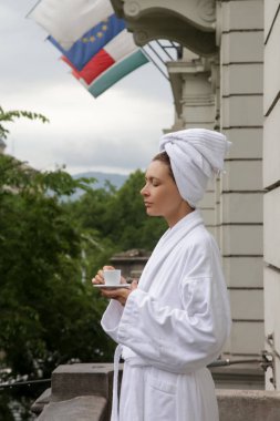 Bornoz ve sarılmış baş havlusu yla balkonda sabah kahvesinin tadını çıkaran kadın