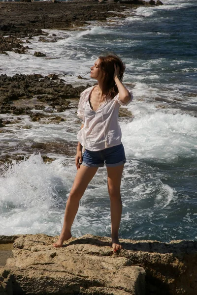 風の強い岩の海岸に白いリネンシャツとデニムショートパンツを着た若いかわいい女性は 観光スポットから遠く離れた場所で夏休みを楽しんでいます — ストック写真