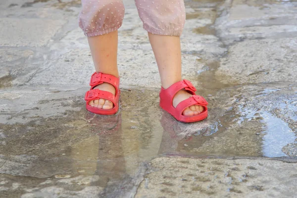 穿着红色橡胶凉鞋的儿童脚跳进雨坑 — 图库照片