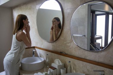 Banyodaki aynanın önünde yüzünü pamukla temizleyen genç bir kadın.