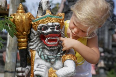 Adorable caucasian toddler girl playing in front of hindu temple. Child playing with traditional colorful barong statue.