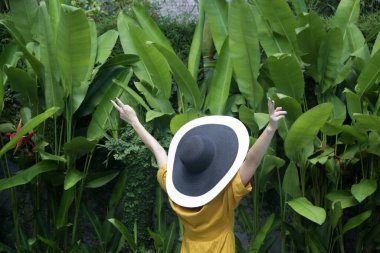 Rear view of woman wearing yellow dress and straw hat in the tropical garden with hands up