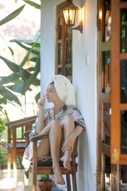 İpek bornozlu ve havluya sarılı genç bir kadın, tahta bir yazlık evde tatilin tadını çıkarıyor. Refah yaşam tarzı konsepti. Idyllic uzak tropikal yaz tatili.