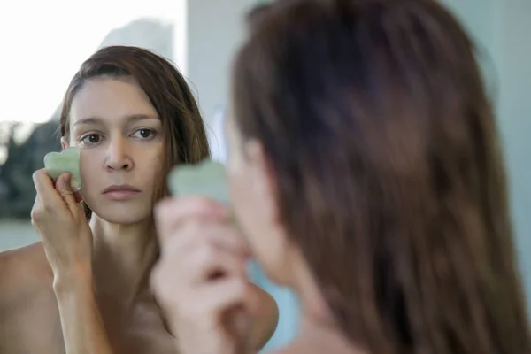 若い女性が鏡の前のバスルームでGuaの沙顔のマッサージをしています 家の治療でGuaの沙玉石 — ストック写真