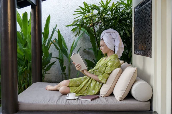 若いですCaucasian女性でローブと髪に包まれたタオルで家に座って本を読んで — ストック写真