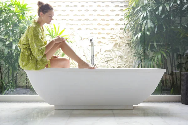 風呂の後に足に保湿剤を塗る浴室の朝の若い女性 — ストック写真