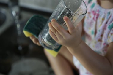 Çocuk mutfakta bulaşıkları yıkıyor. İki yaşındaki tatlı bir kız bardağı lavaboda yıkıyor. Erken gelişim, bebek ebeveynlere yardım eder. Çocuklar için ev işleri.