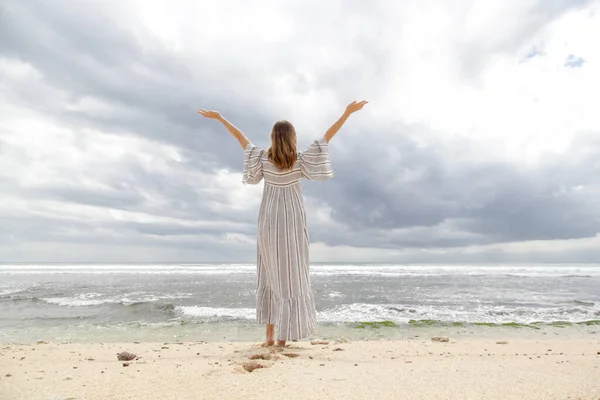 女性は腕を上げ 水平線を見てビーチに立っている 暗い夏の日の間に海岸沿いの長い白いドレスを着た女性のバックビュー 肯定の概念 — ストック写真
