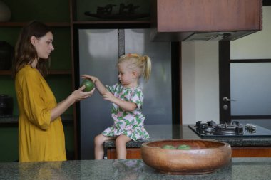 Anne kızına bir elma veriyor. Sevimli küçük kız mutfak tezgahında oturmuş annesiyle vakit geçiriyor..