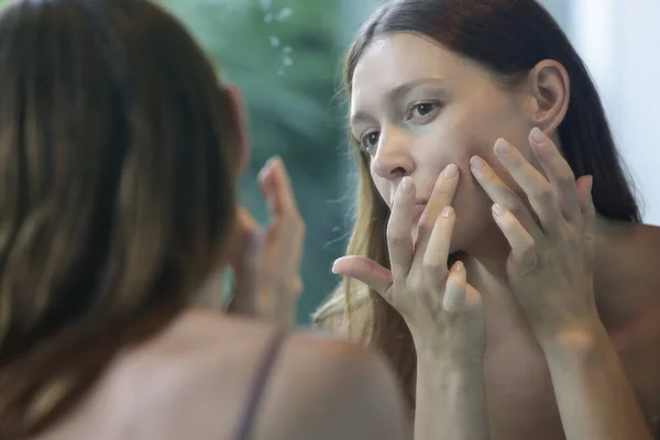 浴室に立つ美しい若い女性の肖像画と鏡の中で彼女の顔を調べる 問題のにきびが発生しやすい皮膚の概念 — ストック写真