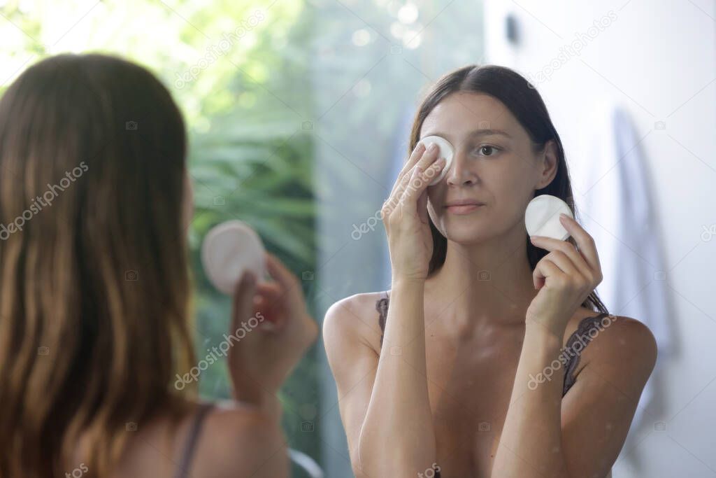 Portrait of a beautiful young woman standing in the bathroom and cleaning her face with cotton pads in front of the mirror, removing makeup, beauty and spa concept.
