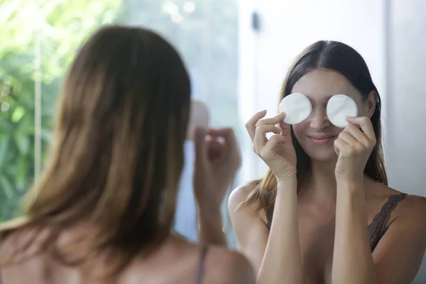 一个漂亮的年轻女子站在浴室里 在镜子前用棉垫擦拭她的脸 去掉妆容 美感和温泉的画像 — 图库照片
